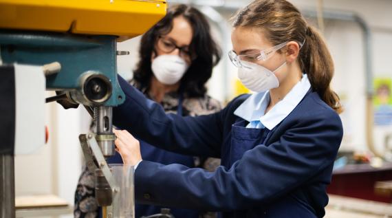 A student in class using the power drill, supervised by a teacher.