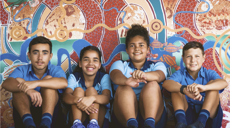 A group of Aboriginal boys in their school uniforms sitting in front of Aboriginal artwork.